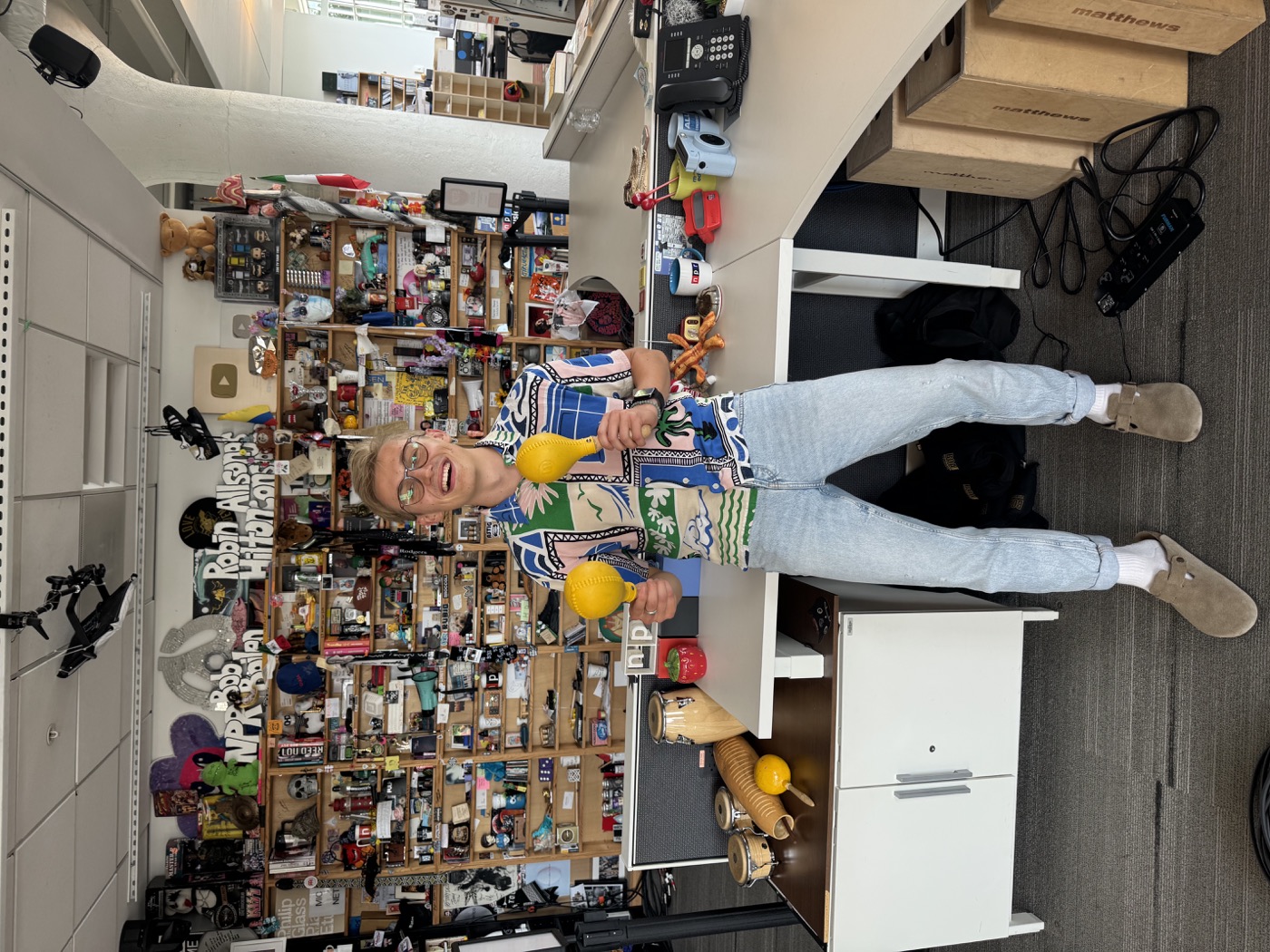 Michal Ruprecht standing in front of NPR’s Tiny Desk holding yellow maracas