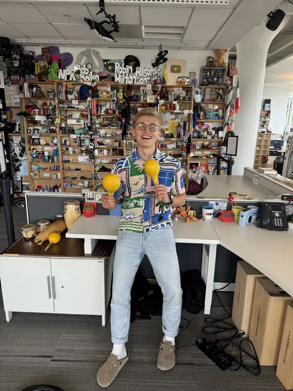 Michal Ruprecht standing in front of NPR’s Tiny Desk holding yellow maracas